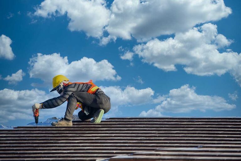 Person inspecting roof shingles beside a ladder while clean gutters and leafy trees surround a house.